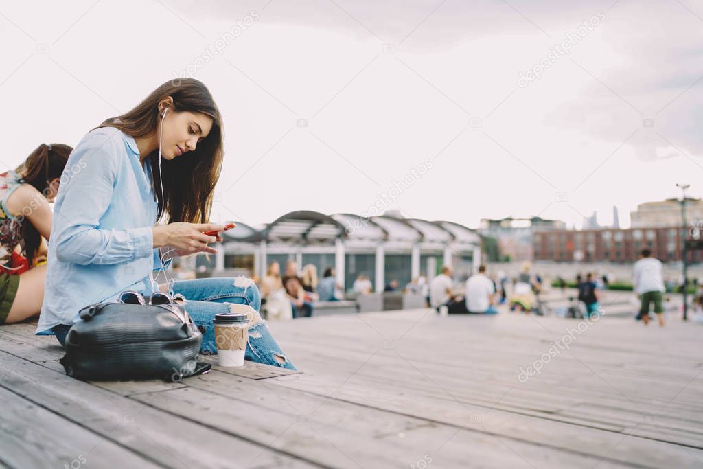 Young hipster girl enjoying coffee break on free time outdoors with favorite music in earphones, millenial woman student listening audio book via smartphone app sitting in college campus on leisure