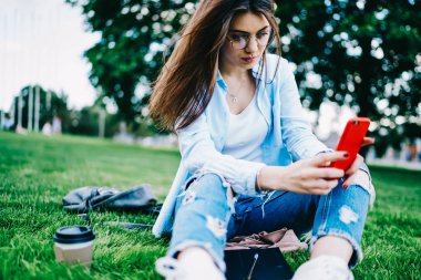 Esmer hippi kız bahar hava, yeşil çimenlerin üzerinde oturan genç kadın cep telefonuyla Park'ta eğlence üzerinde selfie için poz resim yapmak için smartphone kamera kullanarak Eyewear