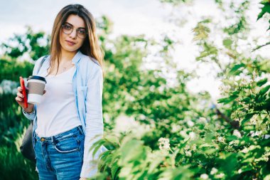 Kahve paket servisi olan restoran ayakta yeşil ağaçlar, yakınındaki serbest zaman üzerinde boş zaman Park kameraya bakarak çekici beyaz kadının yeniden raslantı giyimli hippi kız portresi yarım uzunluk