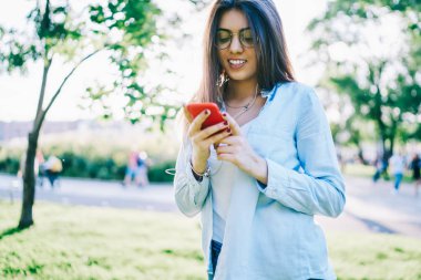 Bahar günü park, harcama yaparken smartphone çalma listesine şarkı İndiriyor Eyewear neşeli hippi kız gülümseyen genç kadın radyo hareket eden telefon ve kulaklık üzerinde boş zaman kullanarak yayını bağlanma