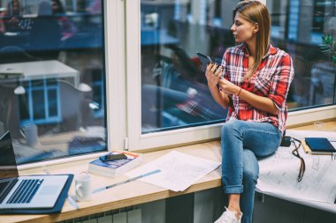 Genç kadın mimar modern dokunmatik yüzey elinde tutan ve kroki bina ile pencere üzerinde oturan Web sitesinde bilgi arama sırasında pencereden dışarı bakıyor rahat kıyafet giymiş