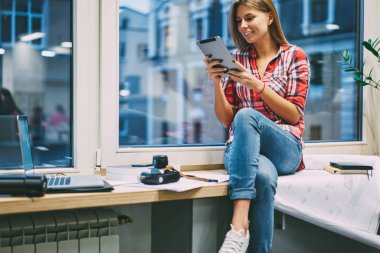 Pozitif genç kadın giyinmiş komik eğitsel bir 4 g kullanarak modern Dokunmatik ped cihaz izlerken rahat kıyafet içinde pencere ofiste oturan internet. Neşeli sarışın öğrenci bilgi tablet okuma