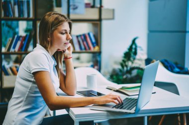 Genç kadın online eğitsel kablosuz internet bağlantısı kullanarak modern Dizüstü aygıtı izlerken konsantre. Dalgın sarışın öğrenci netbook üzerinde App yükleme ve ders çalışma bilgilerini arama