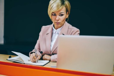 Dalgın deneyimli iş kadını 50 yıl eski izlerken webinar Web sitesinde modern dizüstü bilgisayar bağlı kablosuz 4 g için Internet coworking içinde oturan Not defterinizdeki notlar aşağı yazarken
