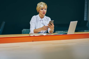 Beyaz gömlekli Smartphone cep telefonu ile gelen bildirim modern dizüstü bilgisayar, iş sırasında okuma emin olgun işkadını. Ciddi kadın profesörü netbook oturup telefon üstünde posta kontrol