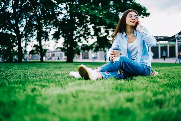 Young hipster girl in casual wear calling to friend on cellular while waiting for meeting in park with coffee to go, young woman spending leisure spring day outdoors having telephone conversation