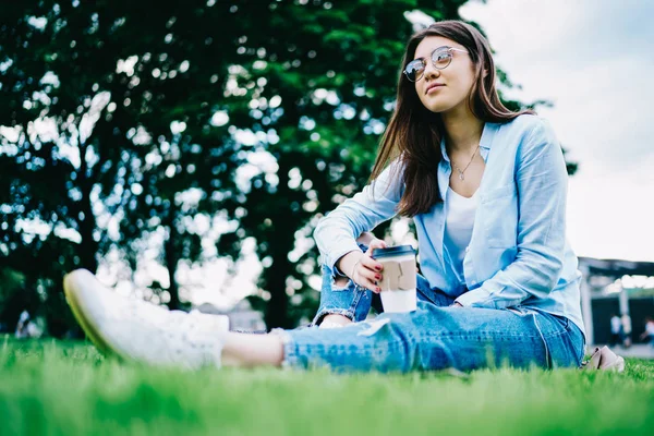 Pensive brunette woman in casual wear sitting on green grass dreaming about something on leisure time, thoughtful female student in spectacles looking away recreating on coffee break in campus