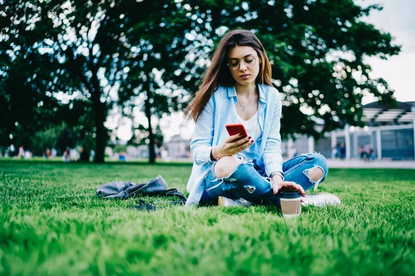 自由時間中に公園で草の上に座ってのスマート フォンで所得のメッセージを読んで眼鏡の若い物思いにふける女性 内気な少女屋外携帯と 接続を使用してソーシャル ネットワークのプロフィールを更新します — ストック写真