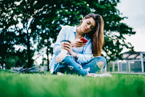 屋外キャンパス チャットの携帯電話経由でコーヒー ブレークを持つ女子学生に草の上に座ってスマート フォン経由で所得メッセージ通知を確認長いブルネットの髪を持つ思いやりのある若い女性 — ストック写真
