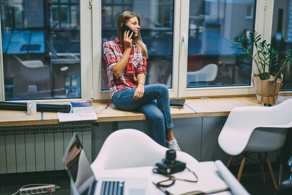欢快的休闲打扮的女设计师呼吁智能手机和笑在电话交谈时坐在窗台上工作休息的办公室 积极的年轻女子谈论细胞 — 图库照片