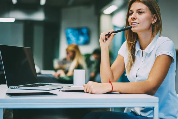 オフィス地区モックアップと現代のラップトップ コンピューターとデスクトップに座ってエッセイを書くための創造的なアイデアを考えながら離れて見て手にペンで思いやりのある笑顔の若い女性 — ストック写真