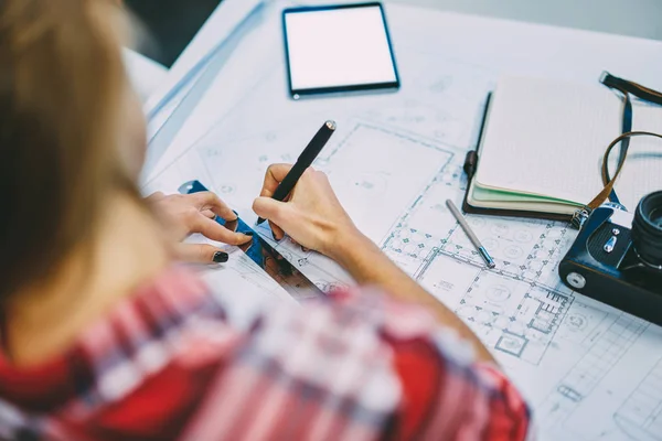 Cropped top view of young woman architect drawing lines on building sketch using ruler and pen.Female engineer writing notes on blueprint sitting at desktop with vintage camera and notepad
