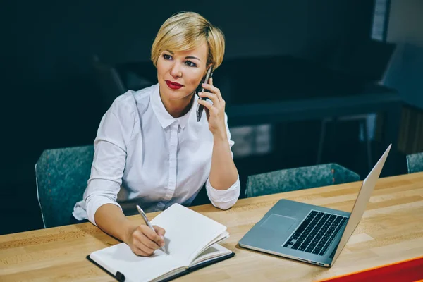 Gorgeous mature female entrepreneur calling with operator on smartphone and writing down text information in notebook working at digital laptop computer.Businesswoman 50 years old talking on cellular