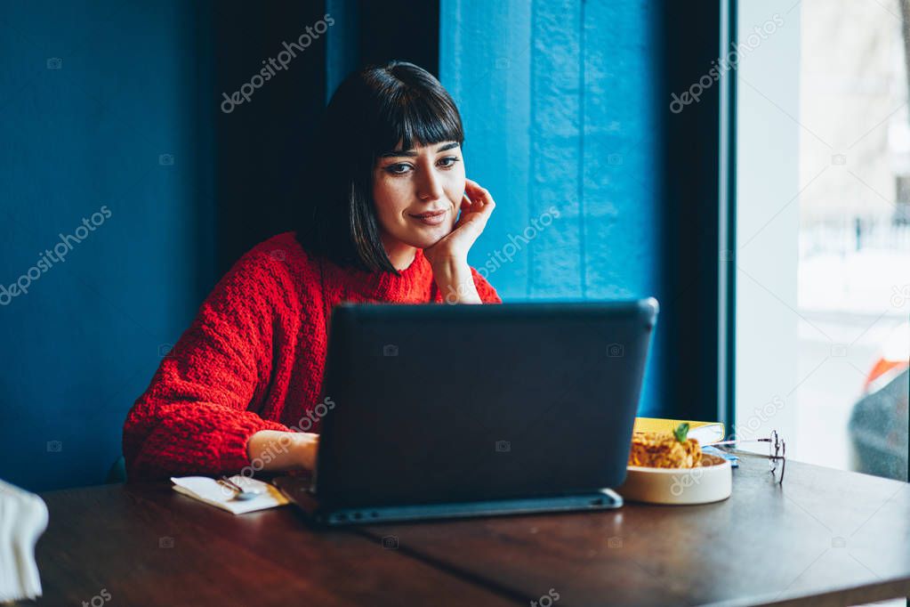 Carefree hipster girl enjoying shopping online via laptop computer during lunch at cafeteria, successful woman watching positive webinar for motivation using netbook and good wifi connection