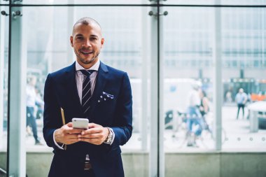 Başarılı genç erkek gurur CEO'su smartphone aygıt ayakta modern ofis üzerinde sms mesaj gönderme portresi. Formal giyim telefon metin yazarken kameraya gülümseyen müreffeh girişimci