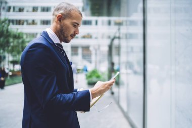 Akıllı telefon elinde tutan ve bildirim okuma finans şirketinin başarılı sahibi yan görünüm. Formal giyim online Caddesi üzerinde hücresel ayakta sohbet müreffeh girişimci