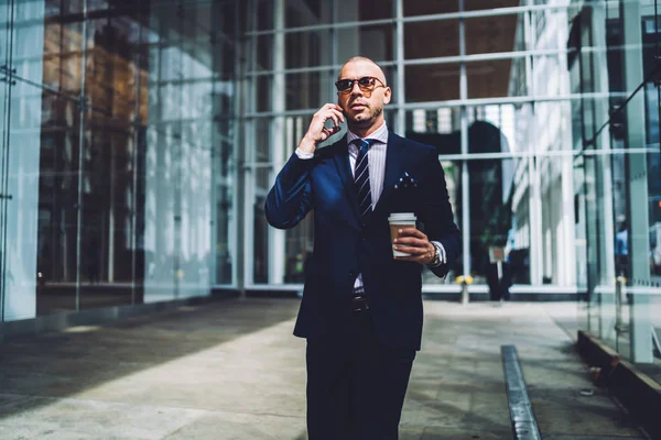 Serious male proud ceo in elegant suit communicating on smartphone during break standing in modern building.Confident busy businessman calling on telephone device while holding coffee to go in hand