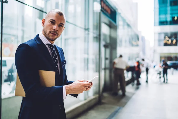 Portrait of confident successful director of financial company looking at camera while downloading app on smartphone using 4G internet.Young businessman in formal wear chatting on telephone