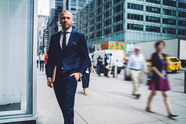 Portrait of confident young entrepreneur dressed in elegant suit holding hand in pocket while looking at camera.Serious businessman with folder for documents standing in urban setting of downtown