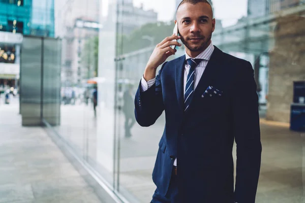 Confident male entrepreneur dressed in formal wear calling on smartphone device and solving business issues standing in urban setting.Serious proud ceo in elegant suit communicating on mobile phone