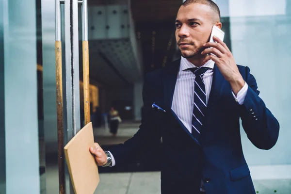 Serious businessman in formal wear going out from office building and communicating during phone conversation on smartphone.Confident young entrepreneur in suit talking on cellular