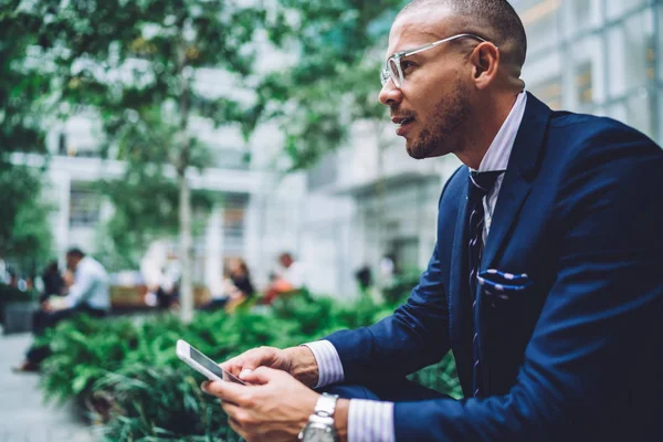 Side view of professional trader in formal wear looking straight while updating app on smartphone using free public 4G internet in urban setting.Businessman sending sms message on mobile phone