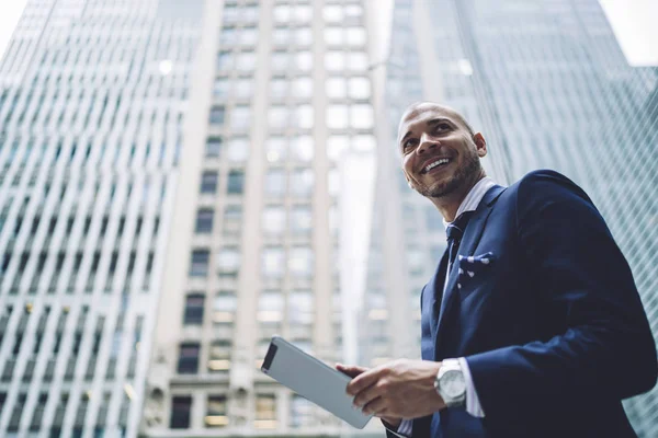 Below view of happy successful entrepreneur dressed in formal wear looking away while installing app on digital tablet using 4G internet walking in modern downtown with high skyscrapers