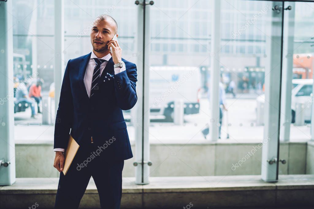 Confident male proud ceo with folder in hand solving business issues during phone call on smartphone.Serious entrepreneur dressed in formal wear talking on digital mobile phone standing in office