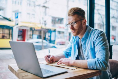 Yetenekli beyaz öğrenci kablosuz internet kullanımı ve kafede oturan notlar iç yapım modern dizüstü bilgisayarda ilginç webinar izliyor. Dalgın genç adam netbook uzaktan çalışma