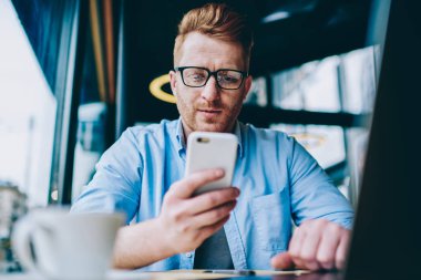 Düşünceli erkek blogger takipçileri Smartphone cep telefonu ile 4 g internet bağlantısı kullanma sosyal ağlar ile sohbet. Hücresel profil güncelleştirme Gözlük Genç adam. Hipster adam cep telefonu haber okuma