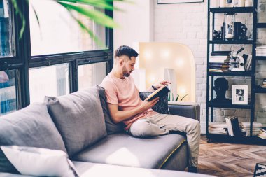 Konsantre hipster adam edebiyat kitap tasarım iç şık apartman dairesinde rahat kanepede oturan konsantre. Daire boş vakit gazetesinin en çok satanlar okuma dalgın genç adam