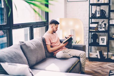 Concentrated hipster guy concentrated on literature book sitting on comfortable couch in stylish flat with design interior.Pensive young man reading bestseller spending leisure time in apartment clipart