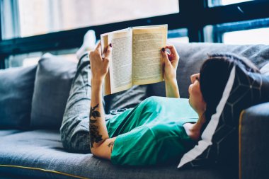 Edebiyat kitap okuma ve rekreasyon vaktinizi rahat kanepede Evde Genç esmer kadın. Modern daire rahat kanepe üzerinde rahatlatıcı elinde ders kitabı ile akıllı hippi kız