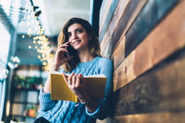 Neşeli hippi kız favori kitap elinde tutan ve olumlu yolu ile smartphone, cep telefonu Üniversitesi Kampüsü üzerinden gülümseyen kadın tebliğ muhabbet ahşap duvarının yakınında ayakta