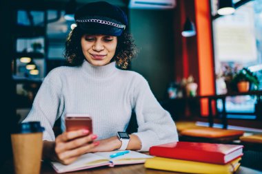 Kontrol için güncelleştirmek üstünde hareket eden aygıt olumlu hippi kız kafeterya wifi için Smartphone vasıl okul sırası çalışma ekipmanları ile oturan mesajlaşma için uygulama yükleme trendy gülümseyen kadın bağlı.