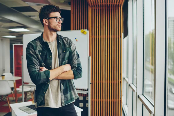 Confident young man in eyeglasses for vision correction looking out of window while thinking on development of own startup project.Serious pondering hipster guy with crossed hands