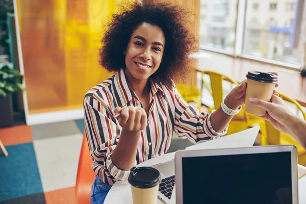 Portrait of african american young woman in casual wear holding coffee to go in hands while smiling at camera and working o freelance at modern laptop computer.Dark skinned female graphic designer