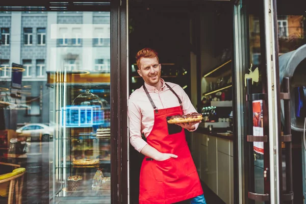 Portrait of happy professional confectioner holding plate with sweet biscuit desserts opening own bakery shop.Successful pastry chef standing at entrance to own cafe with tasty chocolate cakes