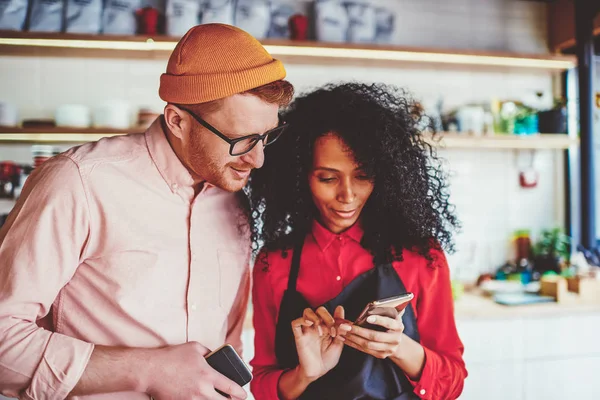 Positive caucasian hipster guy together with cheerful african american waitress watching funny video on website on smartphone using 4G internet.Dark skinned barista showing route on phone hipster guy