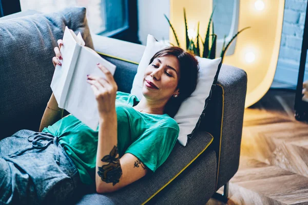 Positive young woman smiling while reading funny story in interesting book enjoying leisure time on weekend at home comfortable interior.Smart hipster girl with bestseller in hand lying on cozy couch