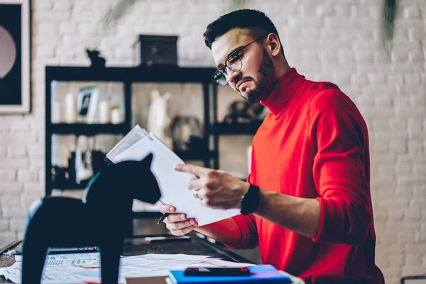 Pensive bearded young man checking course work sitting in modern apartment with design interior.Concentrated hipster guy in eyeglasses reading paper documents with text information at home
