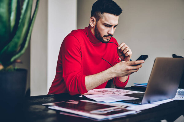 Бдительный молодой человек читает входящие уведомления на смартфоне, работая на фрилансе на современном ноутбуке в домашнем интерьере.
