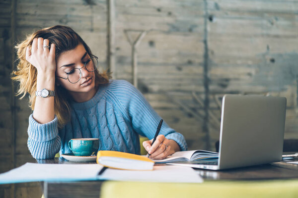 Exhausted female student wear in spectacles for vision correction doing homework in coffee shop reading textbook, tired woman sitting at desktop with book and laptop preparing for college examination