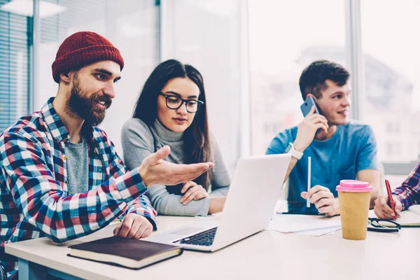 Group of skilled it professionals creating website togetherness using computer, young hipster guy updating software for online work while female colleague sitting next to him at university campu