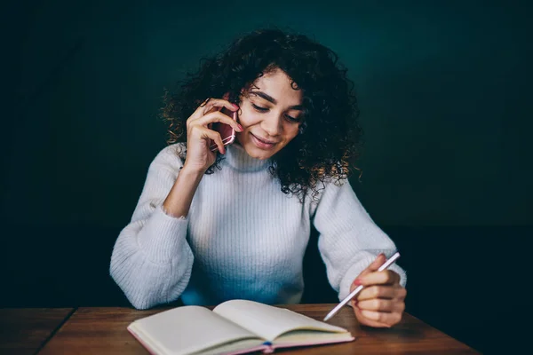 Smiling female journalist making positive conversation talking about time for interview via cellphone, successful hipster girl reading information from textbook during telephone call at campus