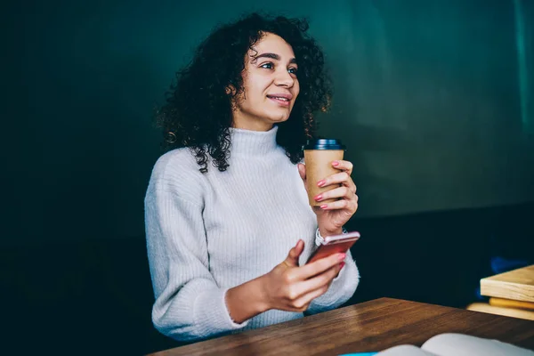 Happy hipster girl looking away holding cup with coffee to go sitting at cafeteria and messaging with friends, millennial positive woman making research for article in social networks via cellphone