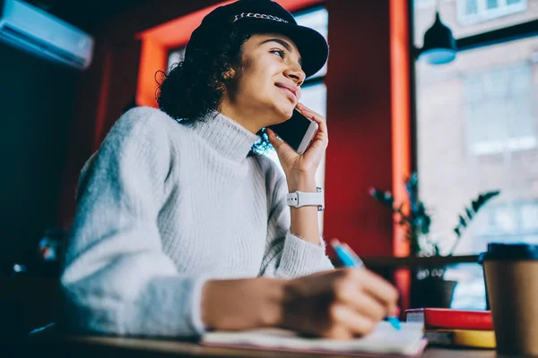 Smiling woman making mobile call connected to wireless while doing homework for school sitting at university cafeteria, positive hipster girl wear in trendy hat and talking with friend via smartphone
