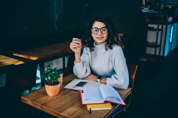 Positive female student preparing for college exam using literature sitting at wooden table, portrait of cheerful hipster girl wear in trendy spectacles for vision correction making research indoors