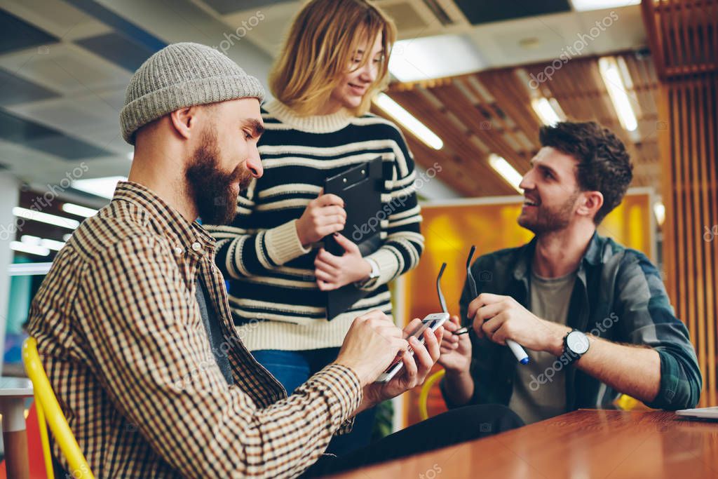 Cheerful male and female colleagues dressed in casual wear communicating with each other during break in modern office.Positive bearded man updating profile on smartphone using 4G internet