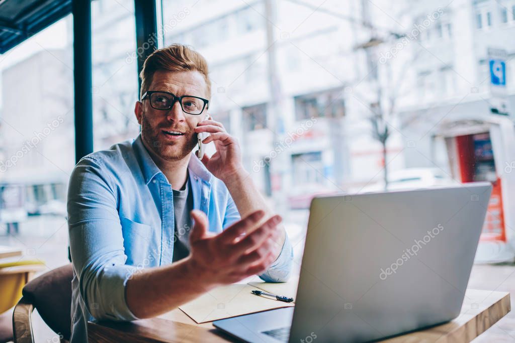 Caucasian young man in casual wear looking away while discussing new tariff plan with operator during phone call on smartphone device sitting at modern laptop computer using wireless internet